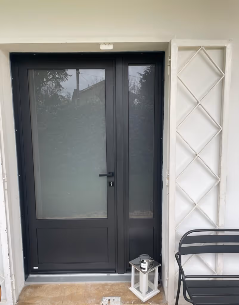 Porte d'entrée moderne en aluminium noir avec une large fenêtre et une lanterne décorative posée au sol à côté d'une chaise noire.