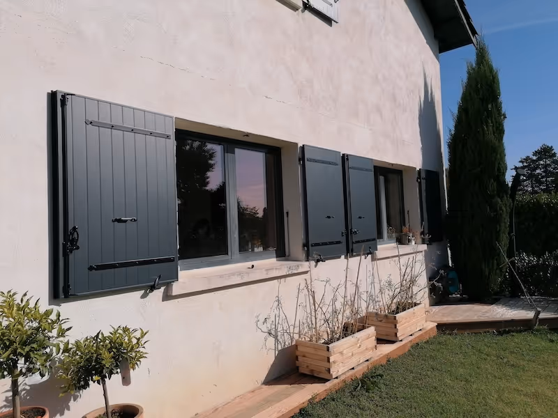 Façade beige d'une maison avec quatre fenêtres équipées de volets noirs, entourée de plantes en pots et jardinières en bois, sur une pelouse verte sous un ciel bleu.