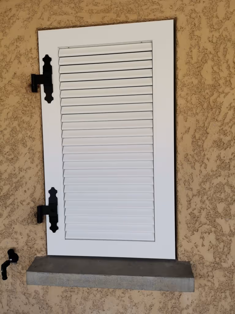 Volet blanc à persiennes fermé avec des charnières noires sur un mur beige texturé et une tablette en béton en dessous.