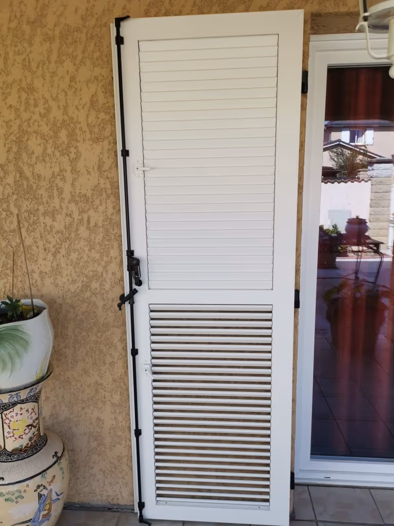 Volet battant blanc fermé avec des persiennes, fixé sur un mur beige à côté d'une porte vitrée et d'un grand pot décoratif.