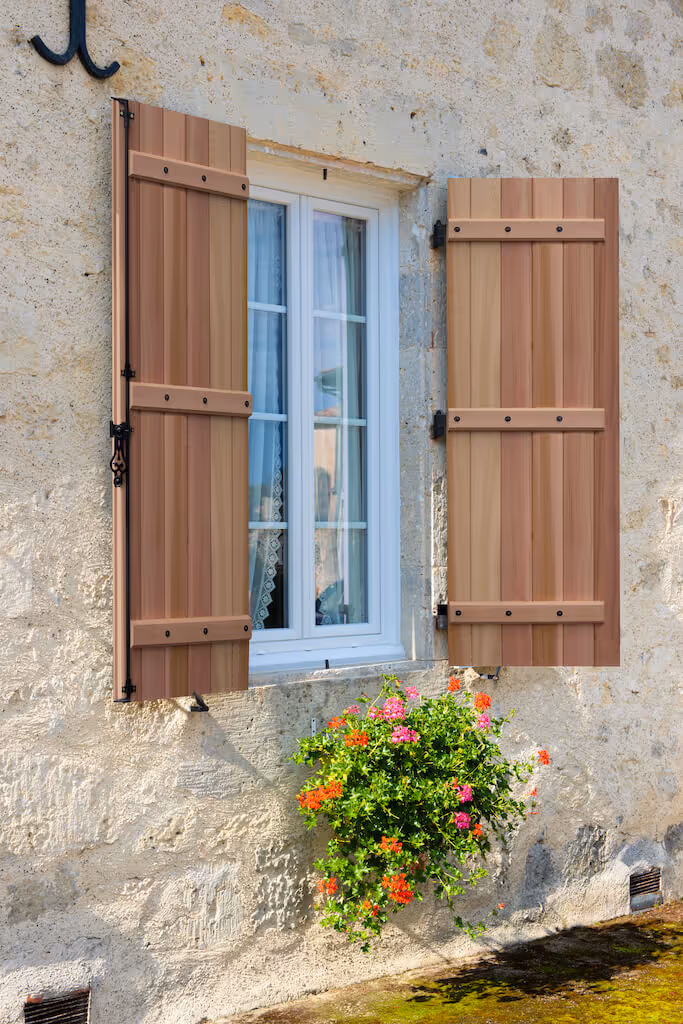 Fenêtre blanche avec volets en bois ouverts sur un mur en pierre beige, avec une jardinière suspendue de fleurs colorées rouges et roses.