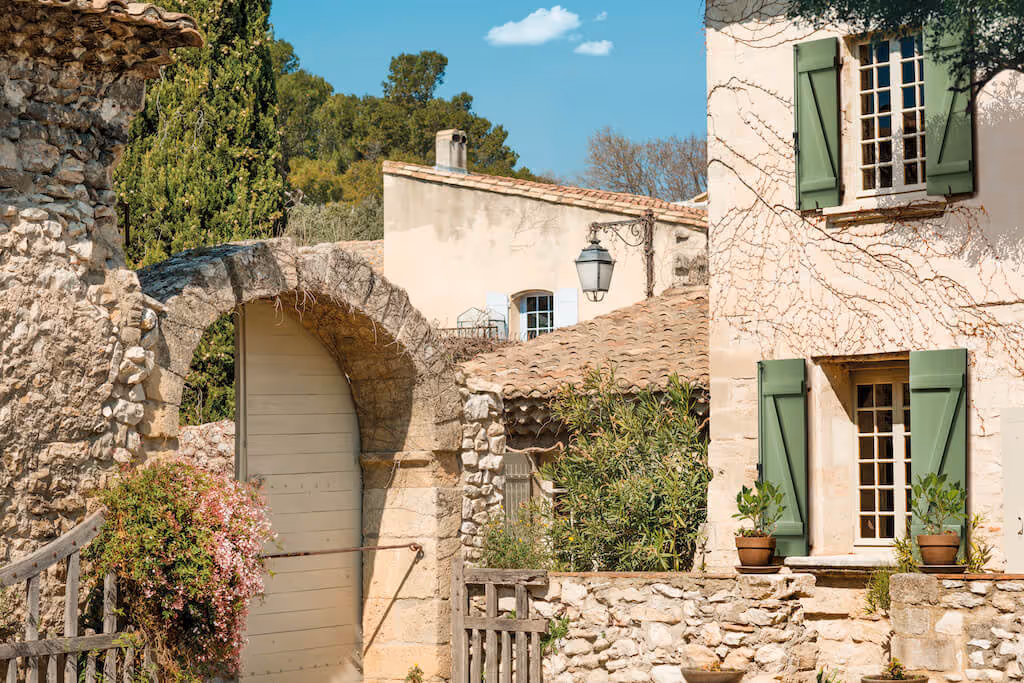Maison en pierre avec volets verts ouverts, porte voûtée en bois clair, plantes en pot et ciel bleu.