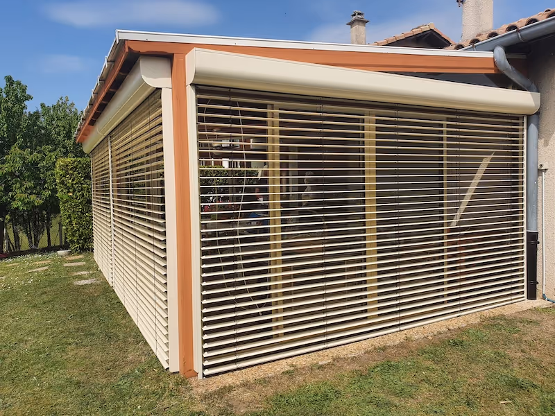 Brise-soleil extérieur en métal beige couvrant une véranda attenante à une maison, avec pelouse et arbre au soleil.