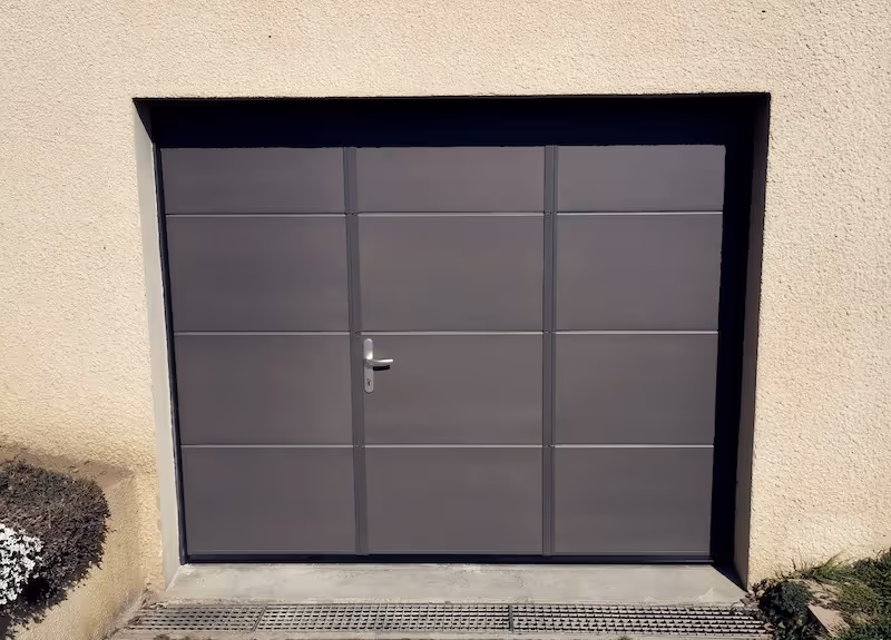 Porte de garage sectionnelle grise avec une porte piétonne intégrée, entourée de murs beige clair.
