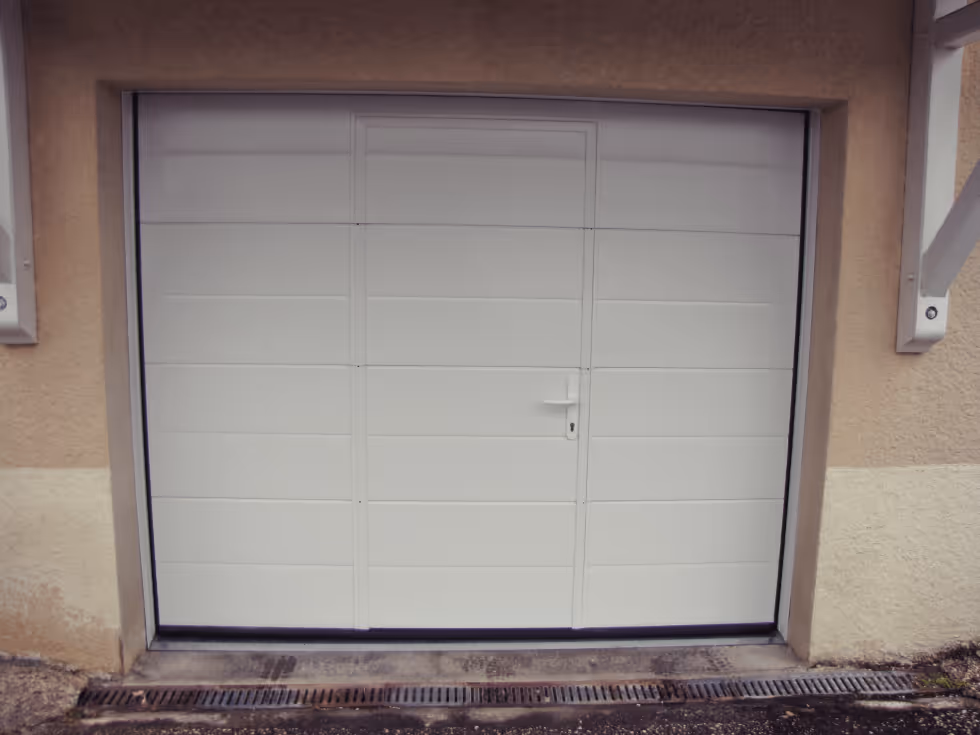 Porte de garage blanche avec une porte piétonne intégrée au centre, encadrée par un mur beige.