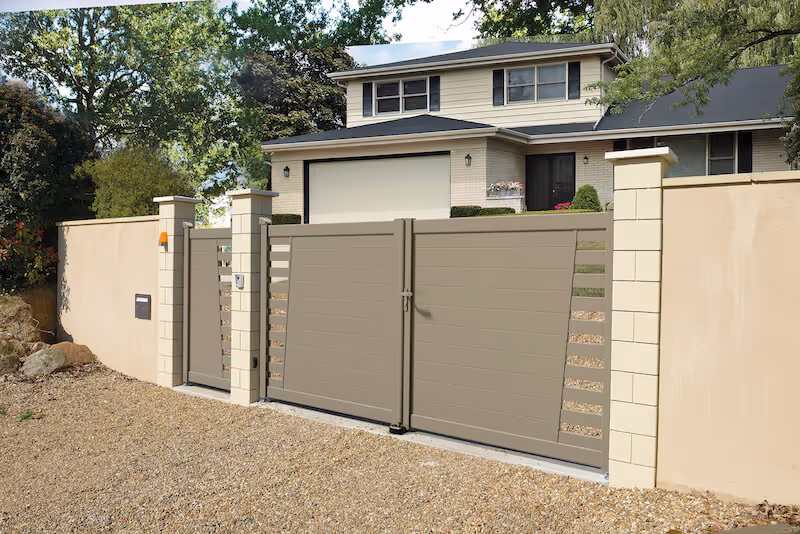 Portail moderne gris avec motifs horizontaux devant une maison blanche à deux étages entourée d'arbres.