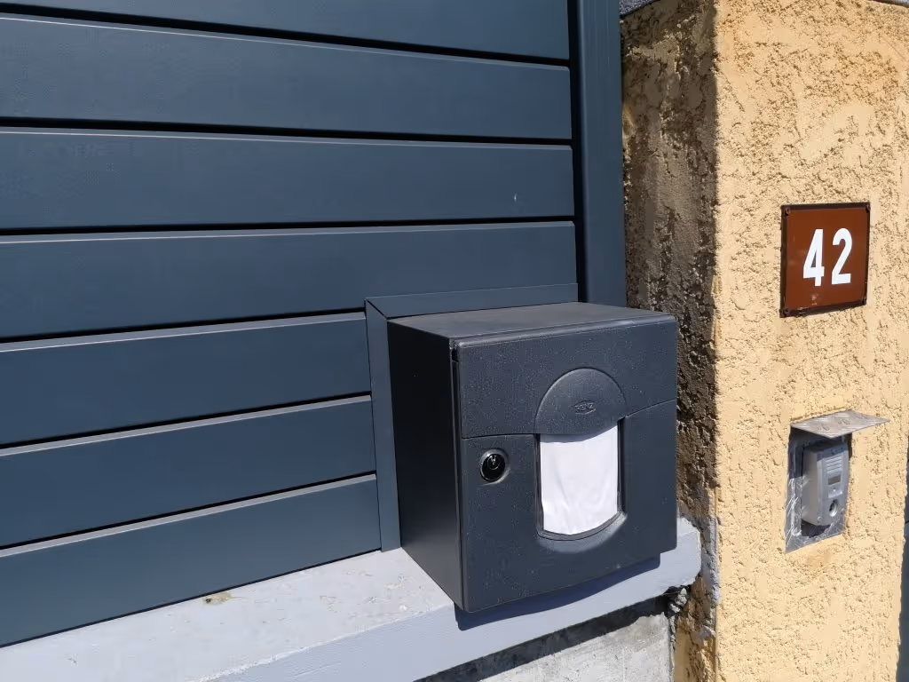 Boîte aux lettres noire montée sur un mur gris à côté d'un mur beige avec une plaque numéro 42 et un interphone.