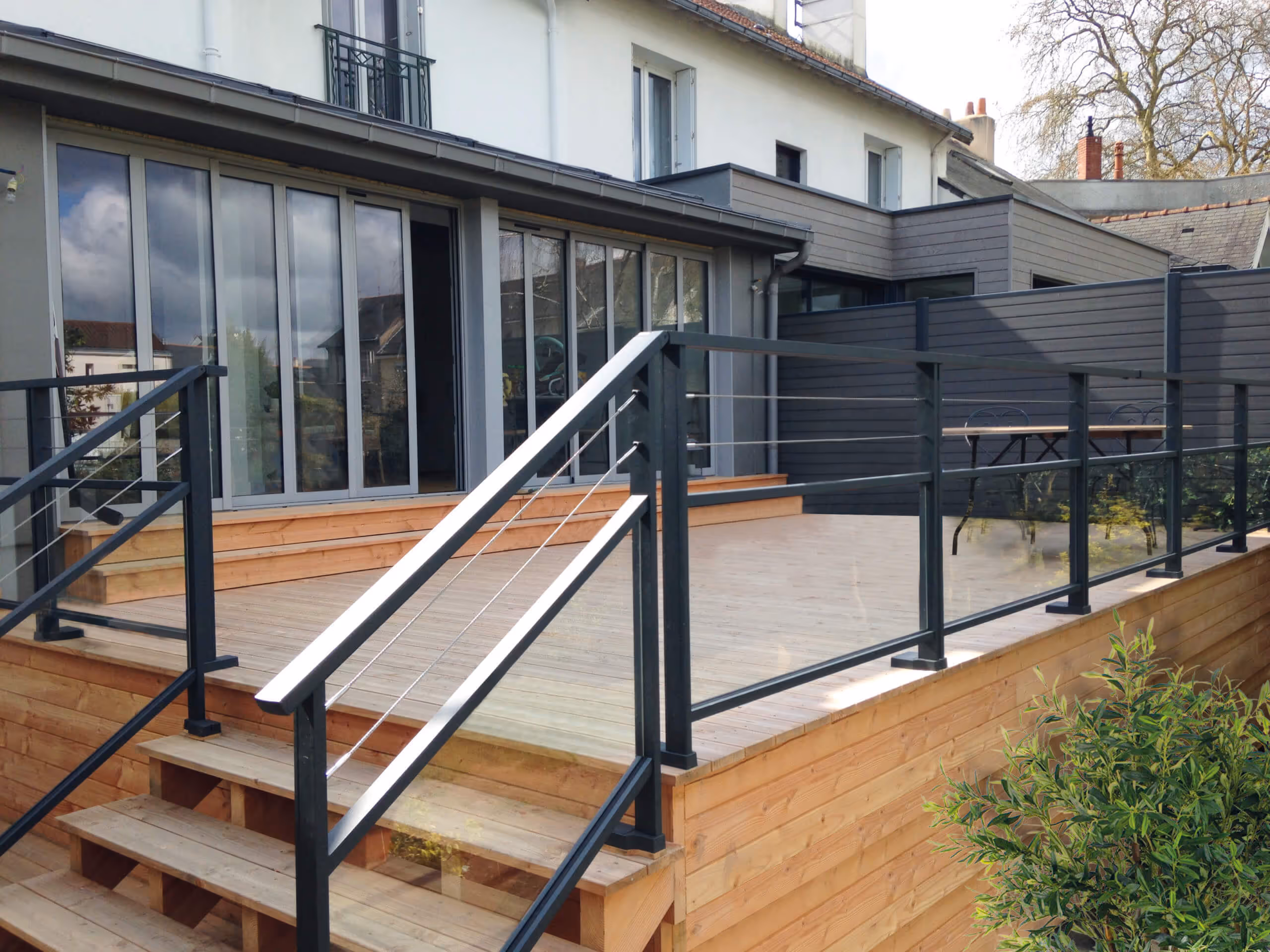 Terrasse en bois avec rambarde noire en métal et verre devant une maison moderne avec grandes baies vitrées.