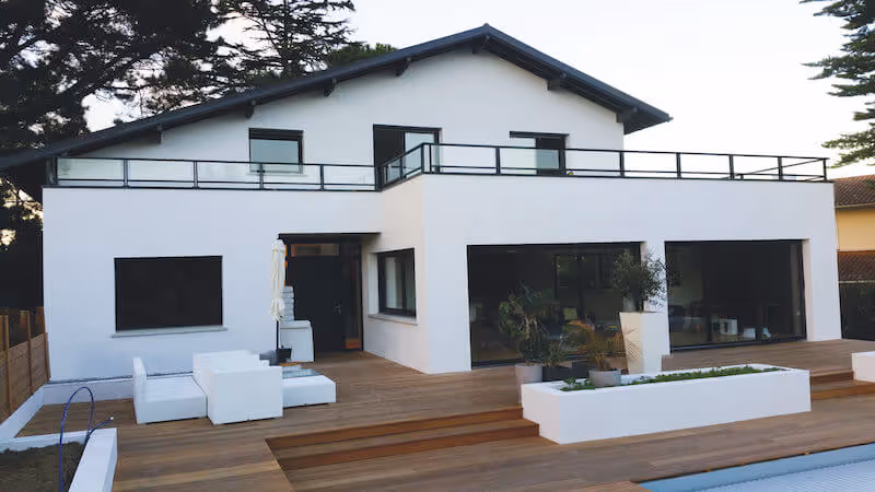Façade arrière d'une maison moderne blanche avec une terrasse en bois, mobilier extérieur blanc et grandes baies vitrées.