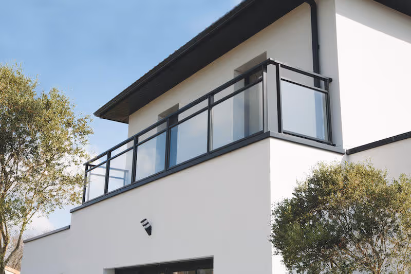 Balcon en verre avec cadre noir sur une maison blanche moderne, entourée d'arbres verts sous un ciel bleu clair.