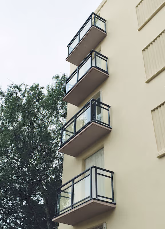 Quatre balcons en verre avec cadre noir alignés verticalement sur un mur beige d'un immeuble, avec un arbre à gauche.