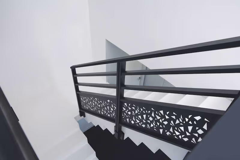 Rampe d'escalier noire avec motif géométrique ajouré, escalier blanc et murs blancs.