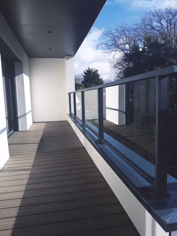 Balcon moderne avec plancher en bois, garde-corps en verre et arbres visibles en arrière-plan.