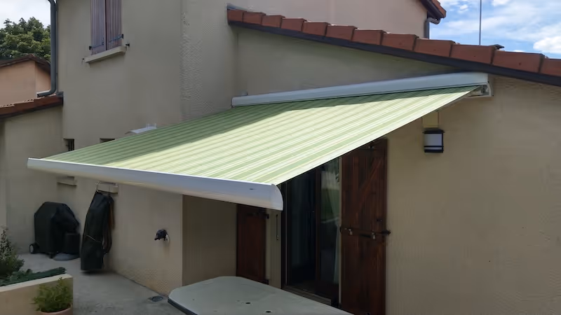Store banne vert clair rayé installé au-dessus d'une porte-fenêtre d'une maison beige avec volets en bois.