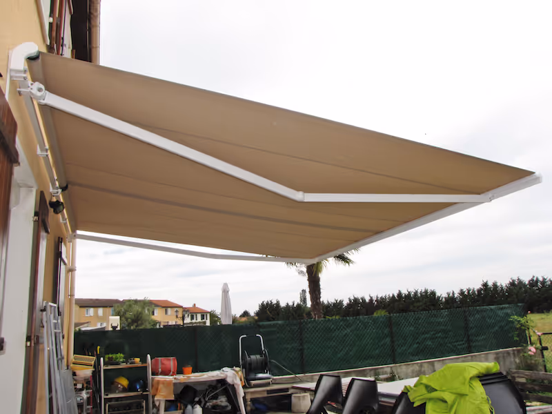 Auvent beige déployé au-dessus d'une terrasse avec des meubles de jardin, entourée d'une haie verte et d'un ciel nuageux.