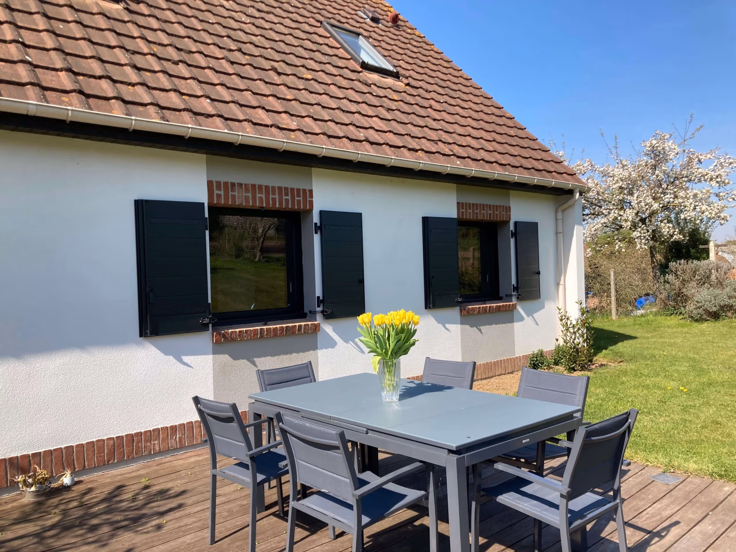 Terrasse en bois avec table grise et six chaises autour, un vase de tulipes jaunes sur la table, maison blanche avec fenêtres à volets noirs, jardin et arbre en fleurs sous un ciel bleu.