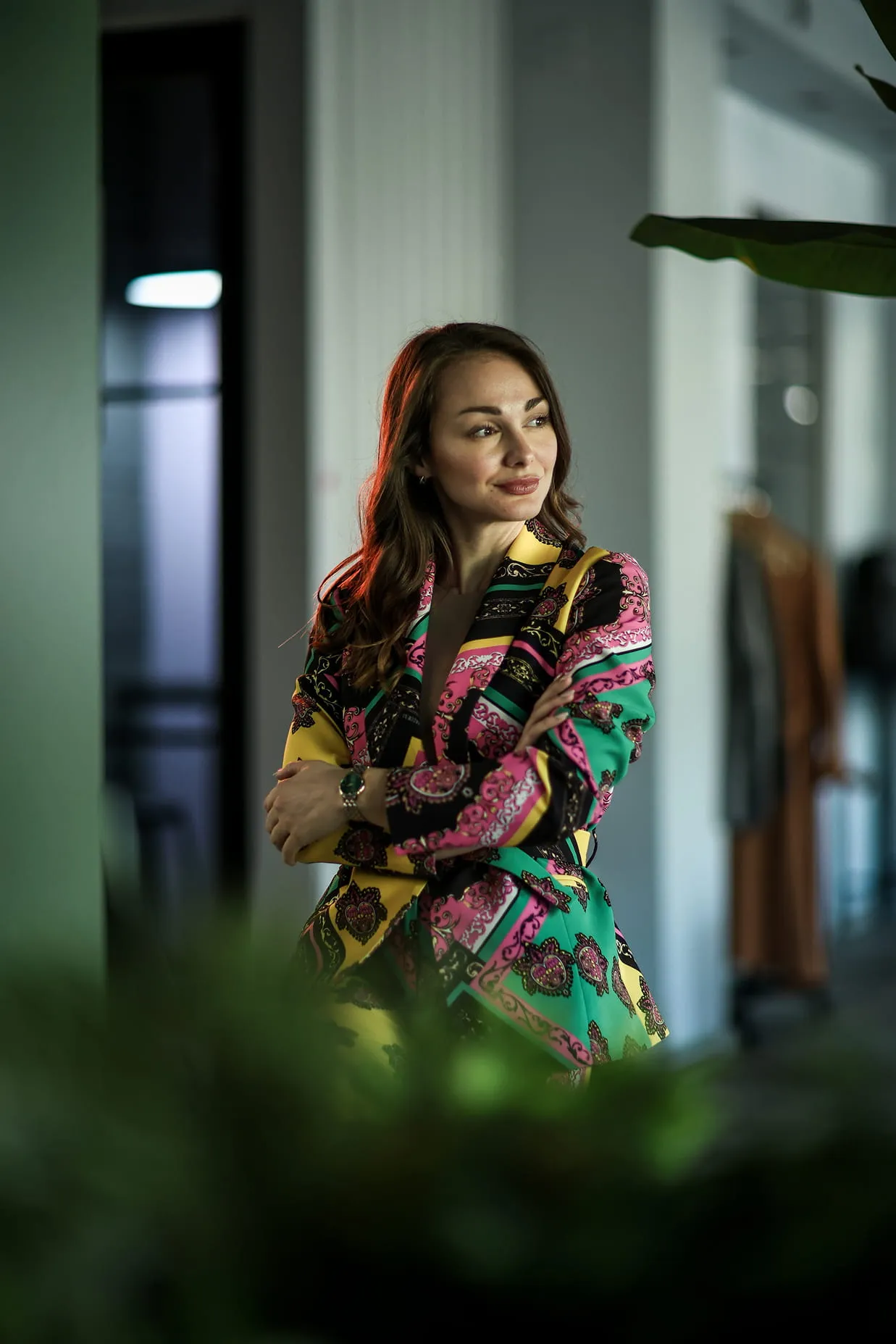 Жена са насмејаним изразом лица у шареном оделу која стоји са преклопљеним рукама у модерном ентеријеру.