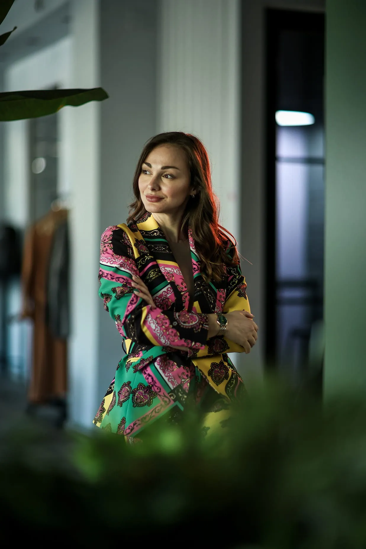 Жена са смеђом косом у шареном оделу, прекрижених руку, у ентеријеру са зеленилом у првом плану.