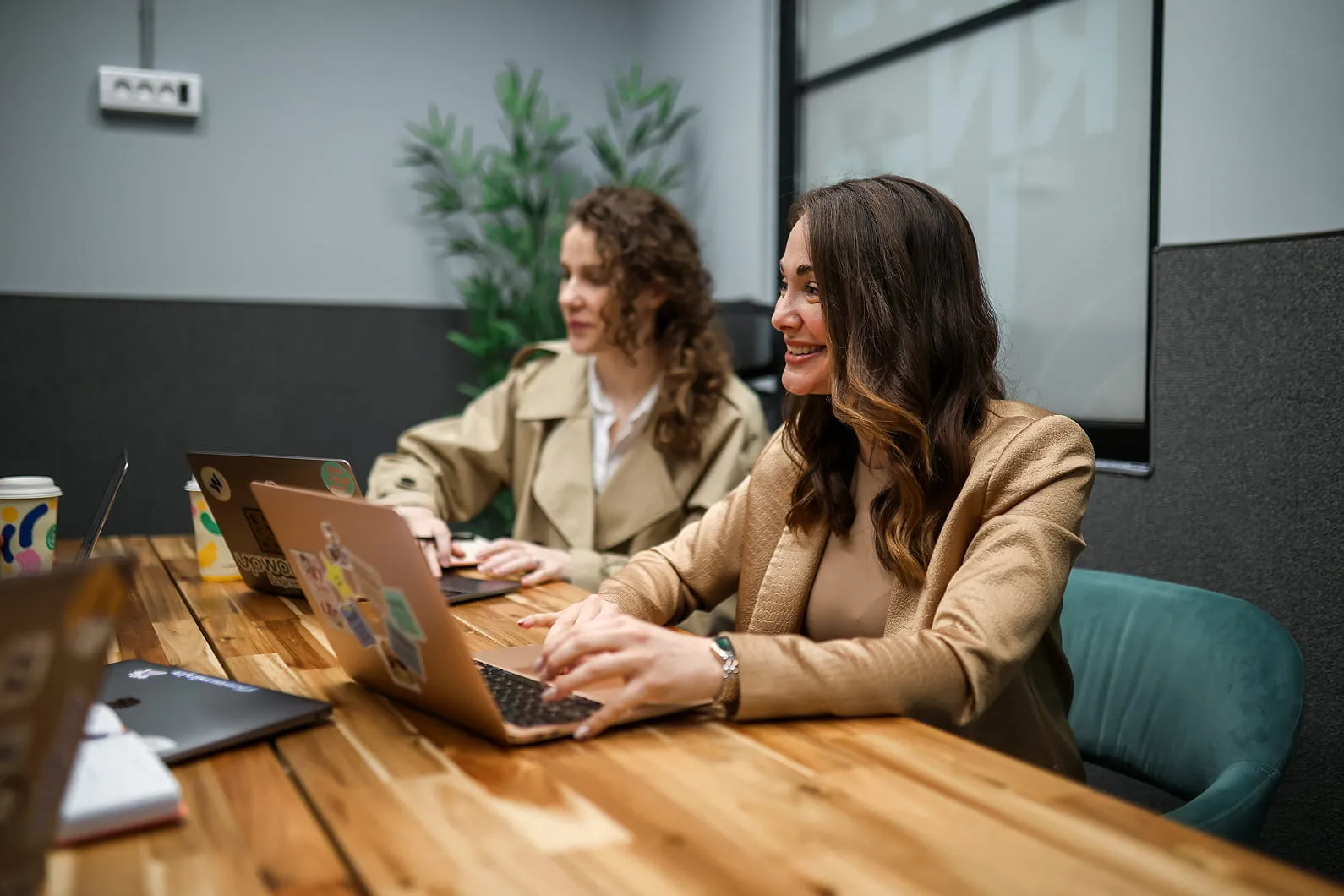 Две жене раде на лаптоповима за дрвеним столом у модерној канцеларији.