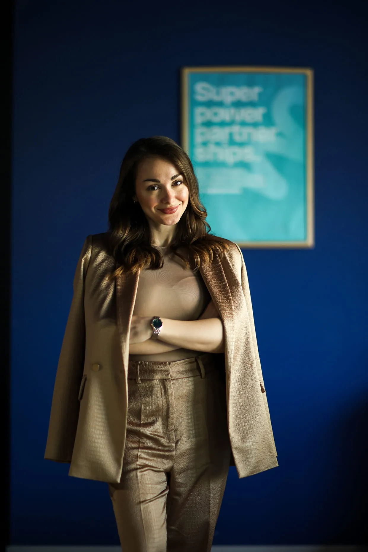 Жена у беж оделу стоји снакрсти руку и смешка се, позадина је плава са нејасним плакатом.