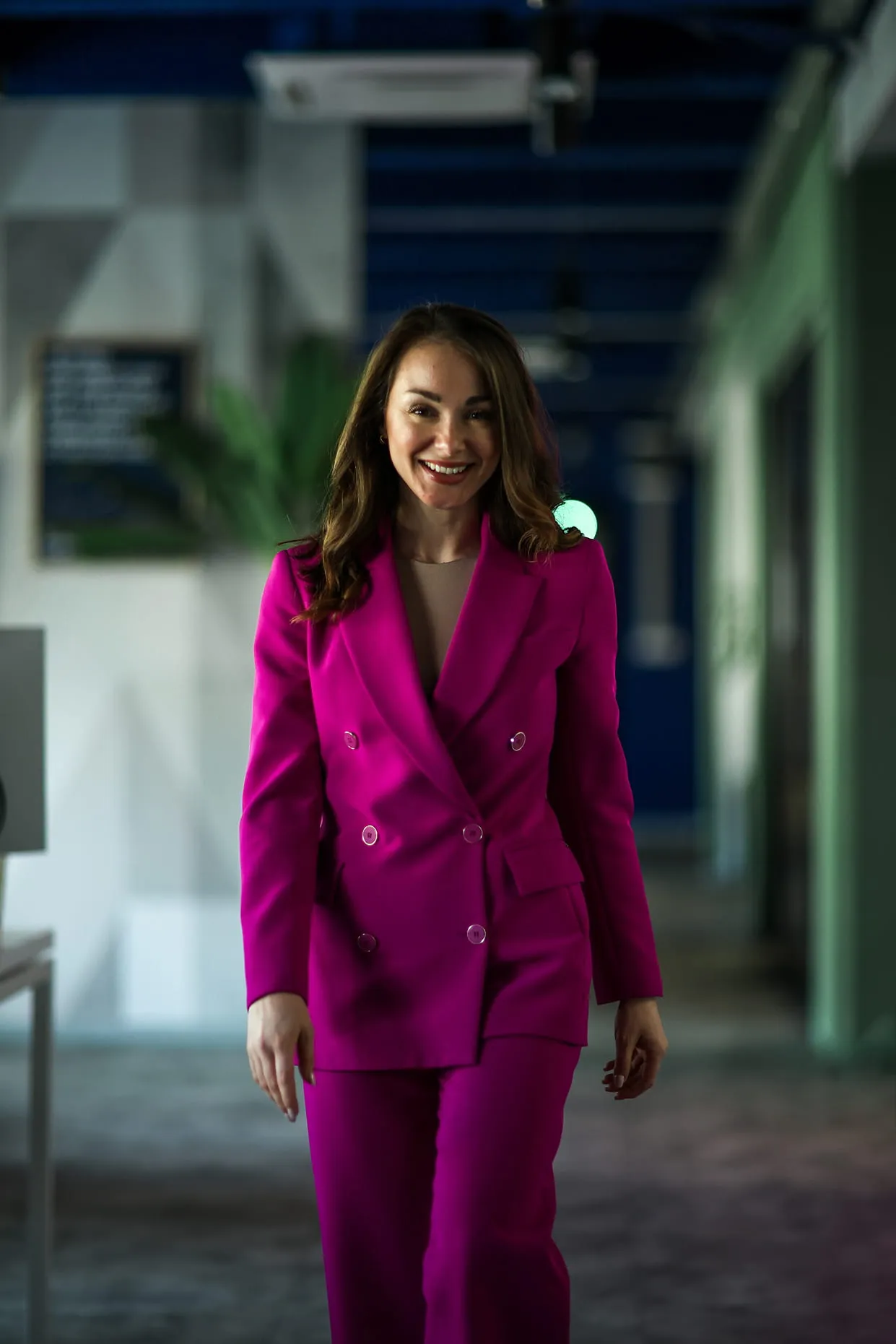 Жена која се смеши у светло ружичастом оделу са дугмадима, шетa пролазом у модерном унутрашњем простору.