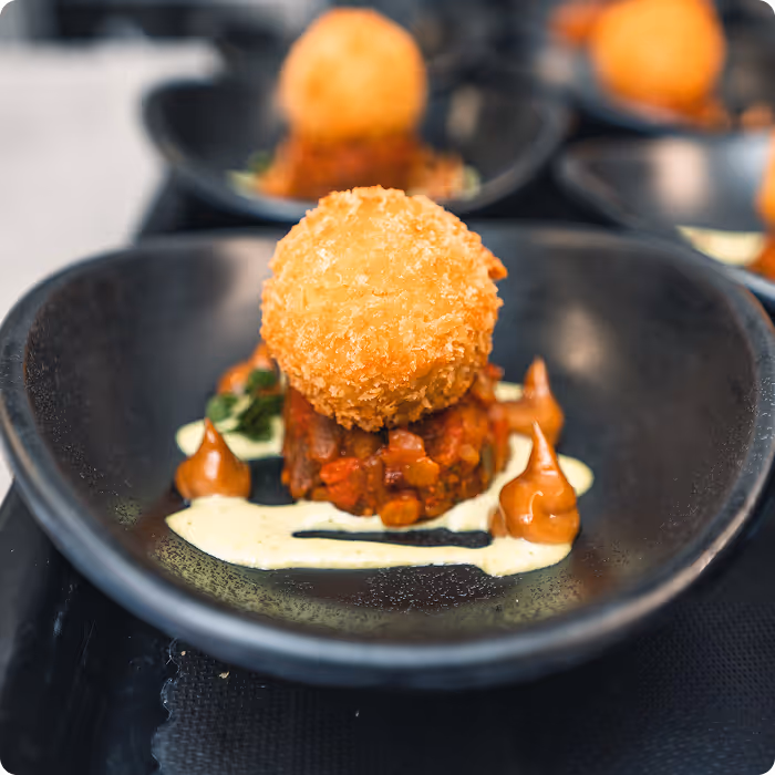 Close-up of a gourmet dish with a breaded fried ball on top of diced vegetables and two sauces artistically arranged on a black plate.
