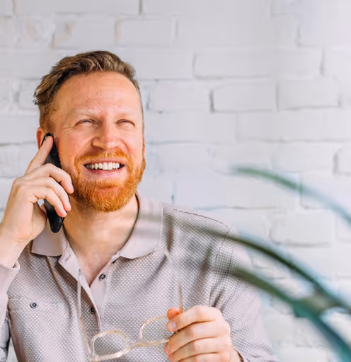 Smiling man with red hair and beard talking on a smartphone while holding eyeglasses, sitting indoors near a white brick wall and green plants.