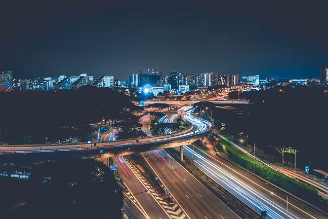 Pont autoroutier incurvé avec des traînées lumineuses laissées par des véhicules en mouvement la nuit dans une zone urbaine avec des bâtiments en arrière-plan.