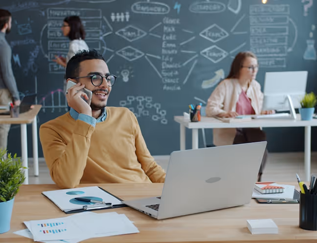 Diverse office workers wearing headsets, collaborating and working on laptops in a modern open-plan office with glass walls and a server rack.
