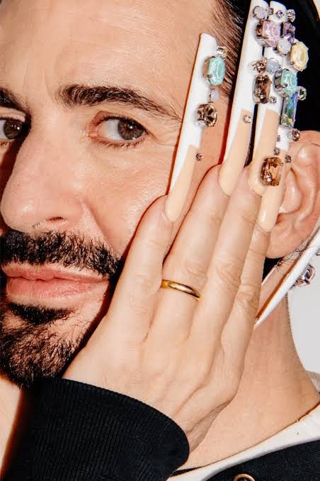 Detail of a hand with very long white nails set with coloured stones.