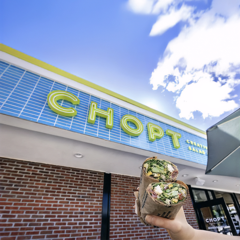 Looking up at the CHOPT sign on its blue tile fascia, a hand holding two wraps in the foreground.