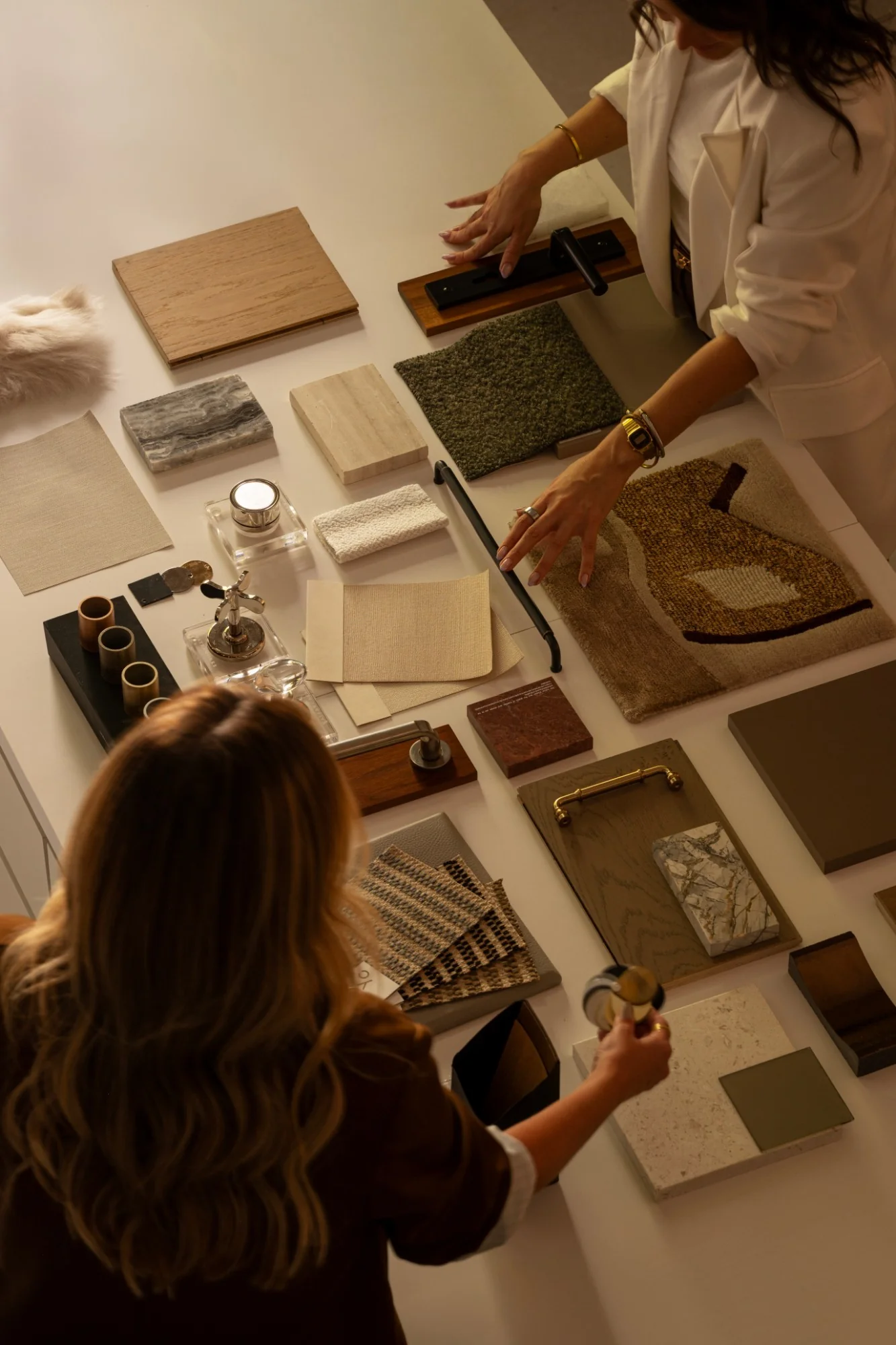 Two interior designers reviewing a table of material samples, including wood veneers, marble and stone tiles, textured fabrics, a hand-tufted rug swatch, and brass and bronze hardware.