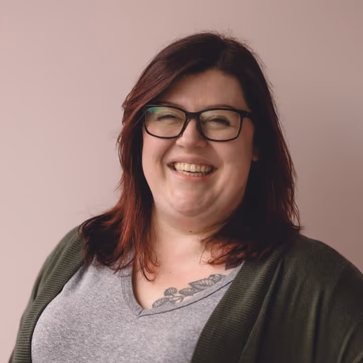 This is a photo of Anne Anderle. Smiling woman with glasses, red hair, and a visible chest tattoo, wearing a grey shirt and green cardigan.