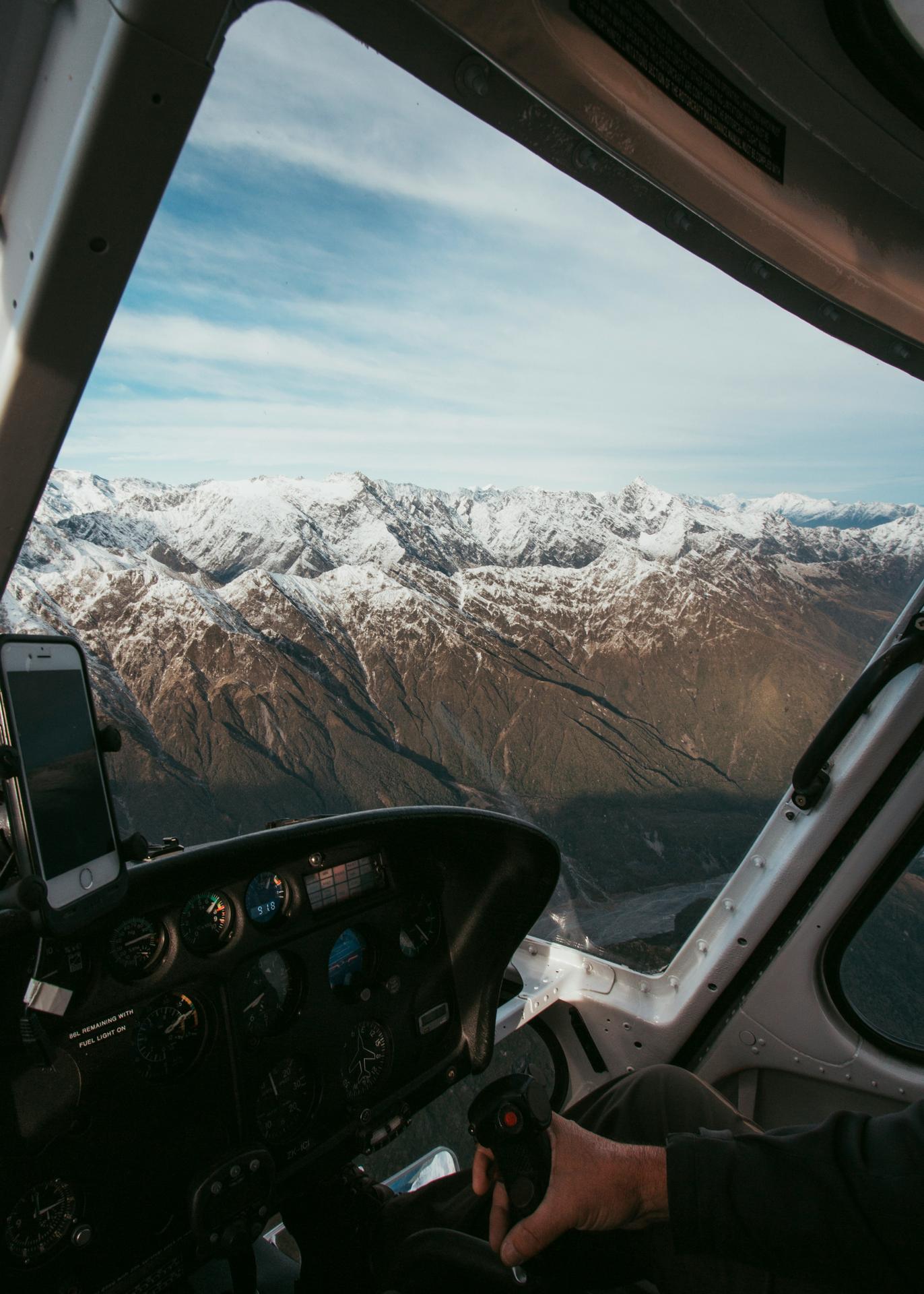 View from a small airplane cockpit showing the pilot's hand on the control stick and snow-capped mountains outside the window.