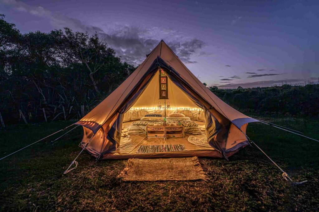 Illuminated glamping tent at twilight with cozy bedding, rugs, and decorative string lights inside.
