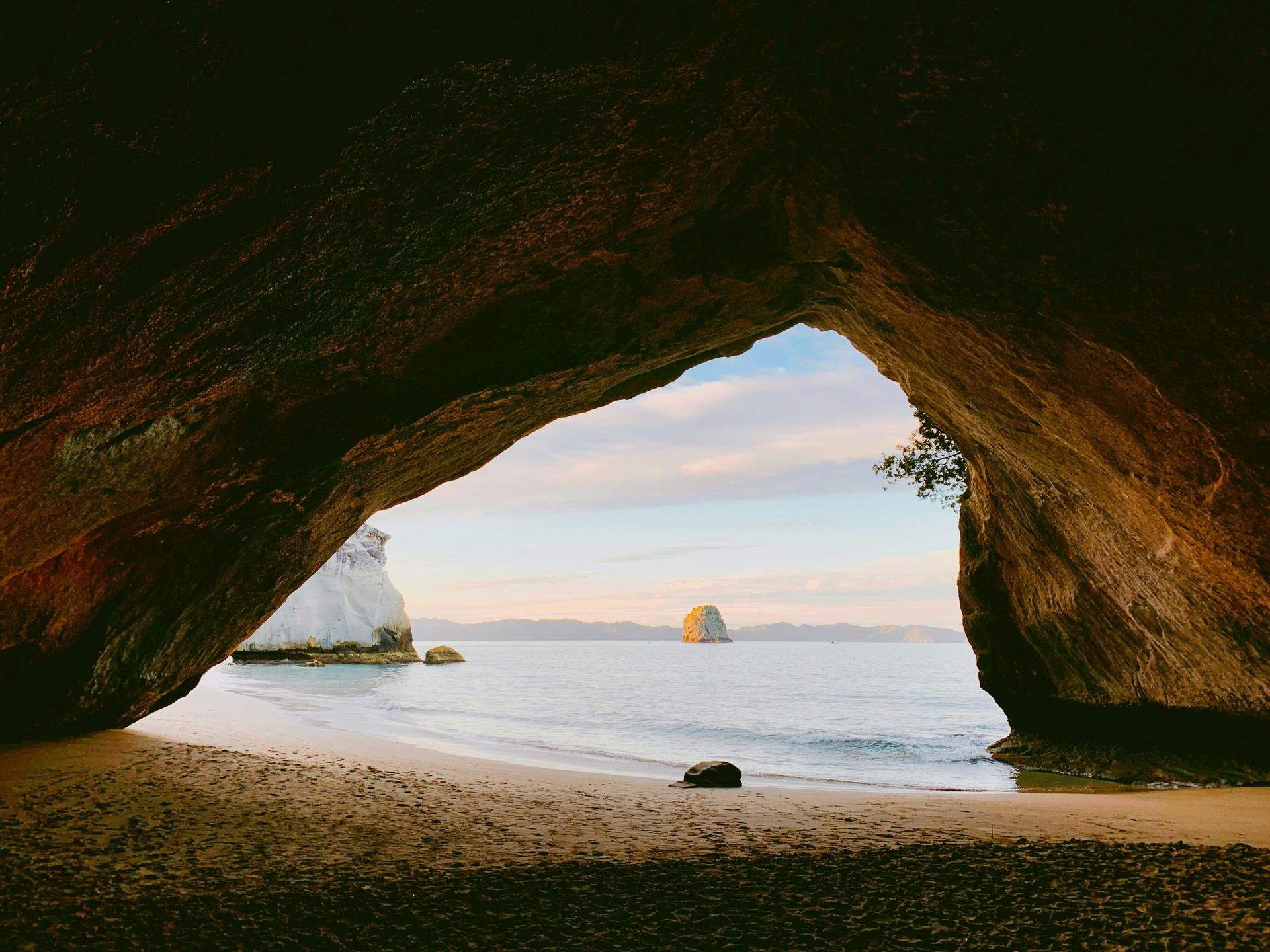 View from inside a large cave looking out to a sandy beach, calm sea, and rocky islands in the distance under a partly cloudy sky.
