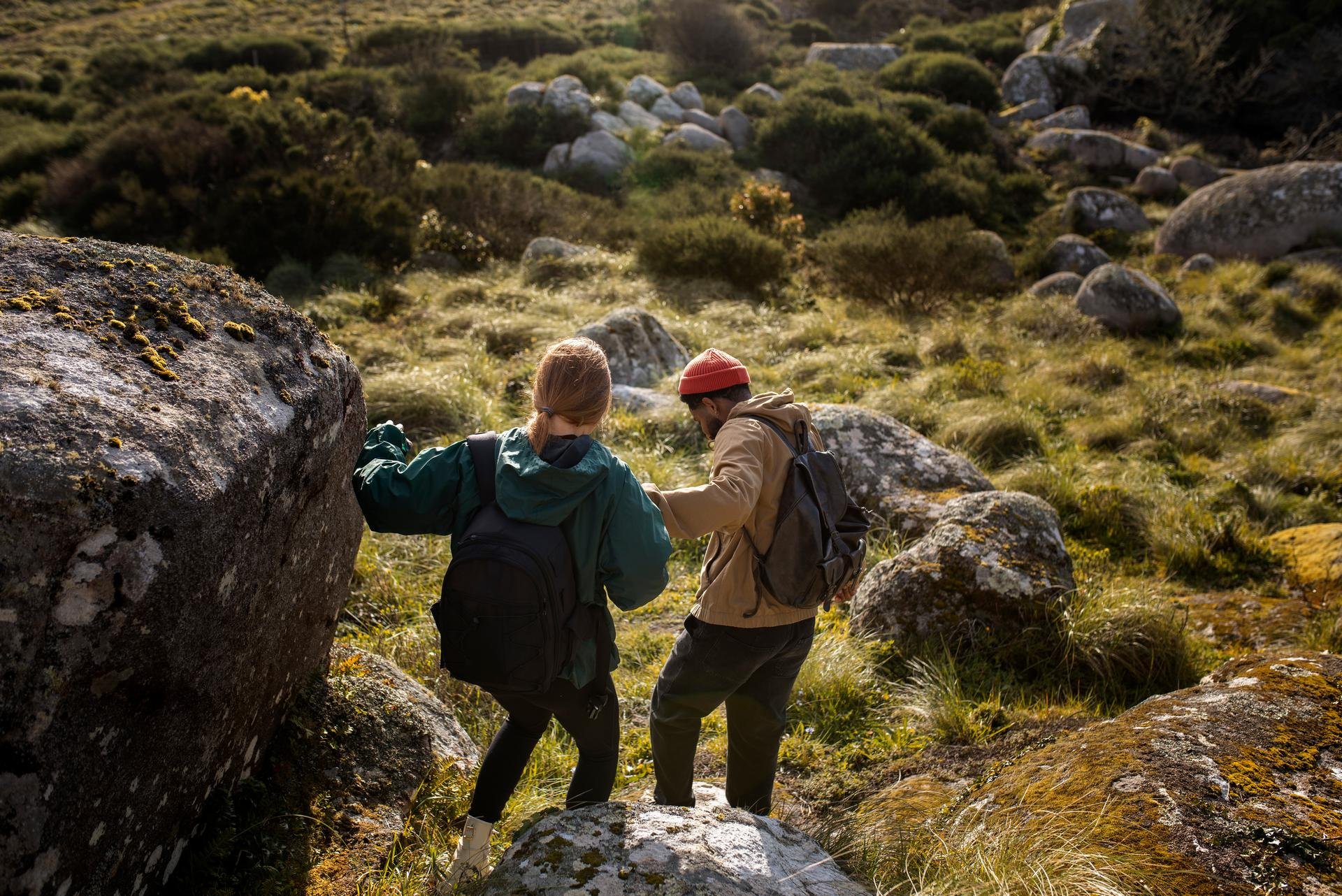 Two hikers with backpacks navigating rocky, grassy terrain in a sunlit natural landscape.