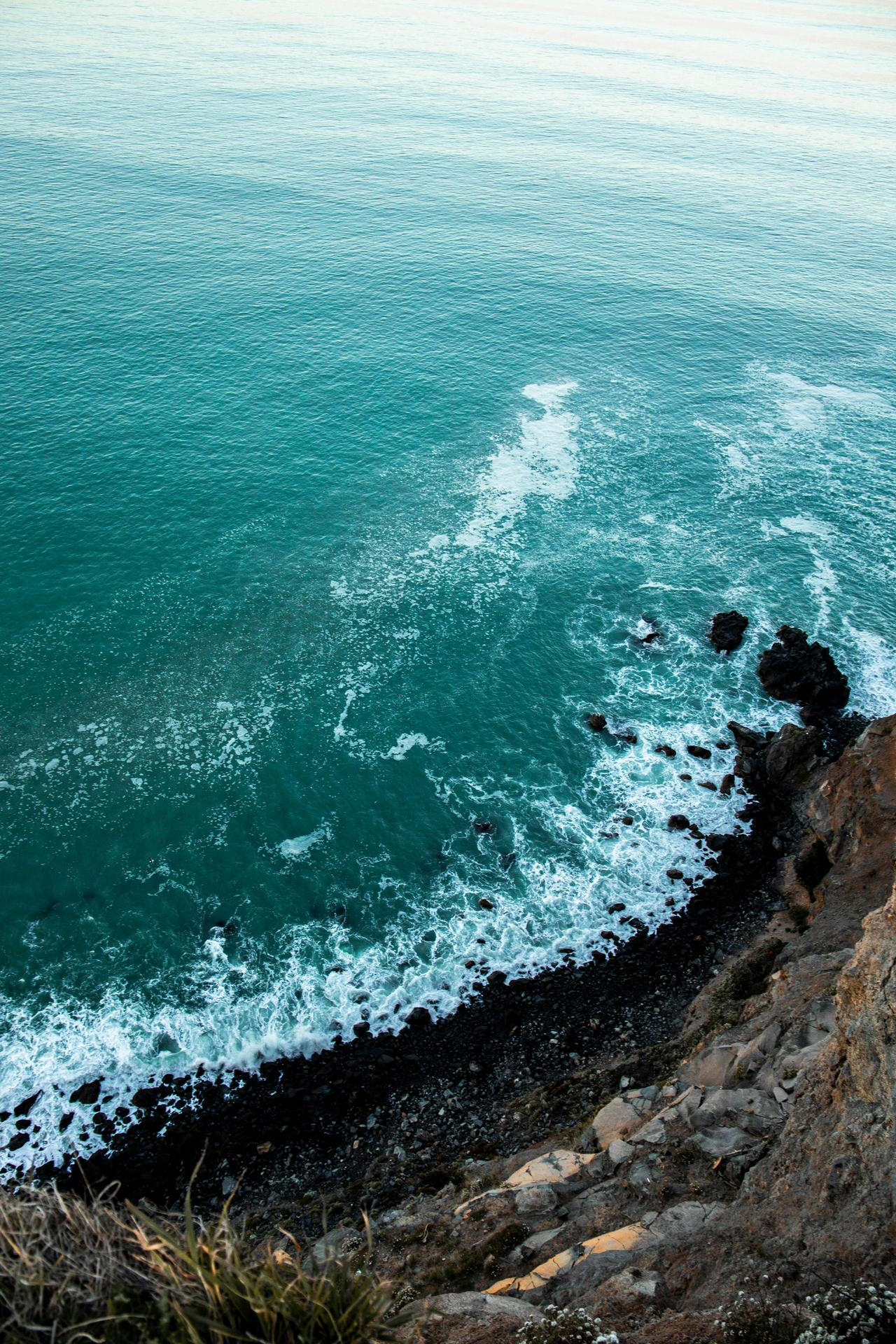 View downward from a rocky cliff to turquoise ocean waves crashing on a dark rocky shoreline.