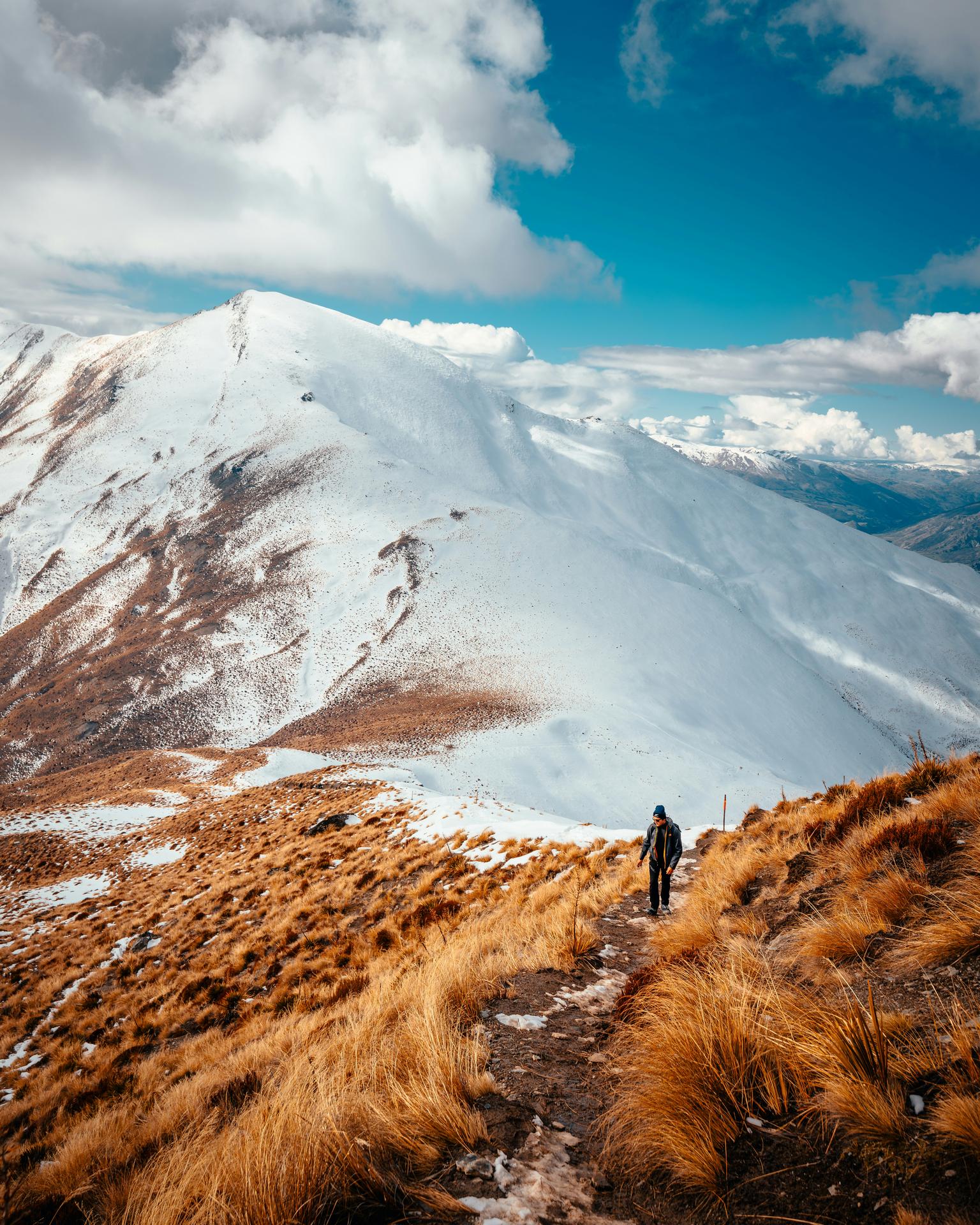 Hiker walking on a narrow mountain path with golden grass and snow-covered peaks under a partly cloudy blue sky.