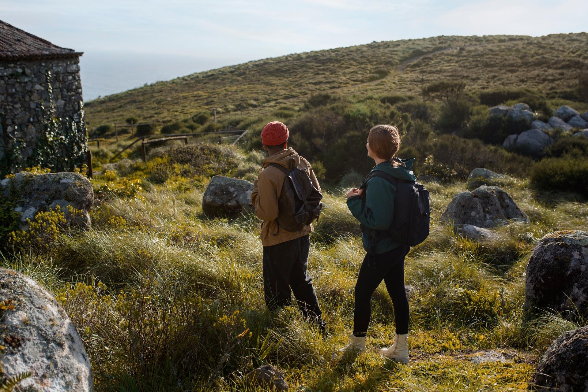 Two hikers with backpacks standing in a grassy field near rocks and an old stone building, overlooking a hilly landscape.