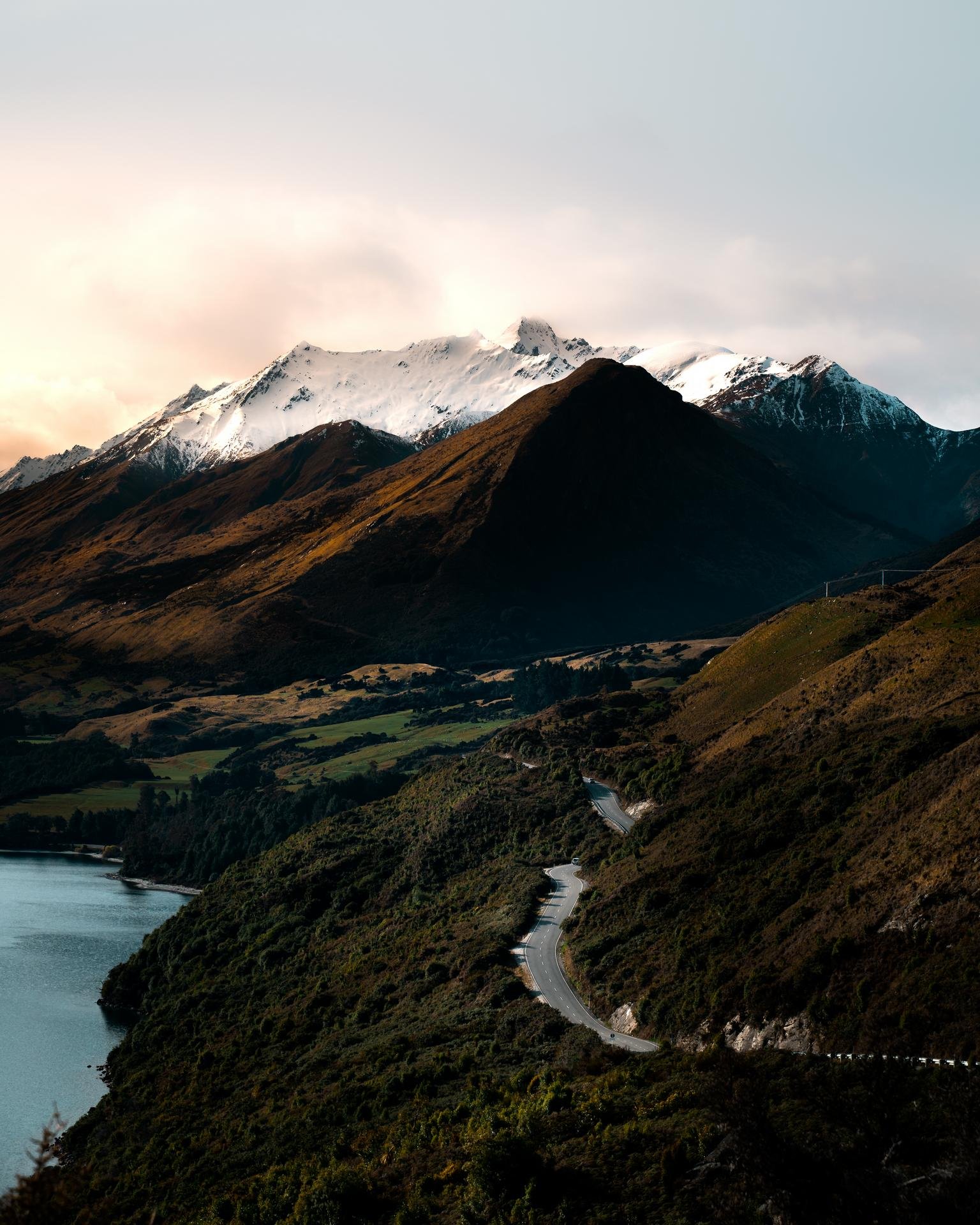 Winding road along a lake with snow-capped mountains in the background at sunset.