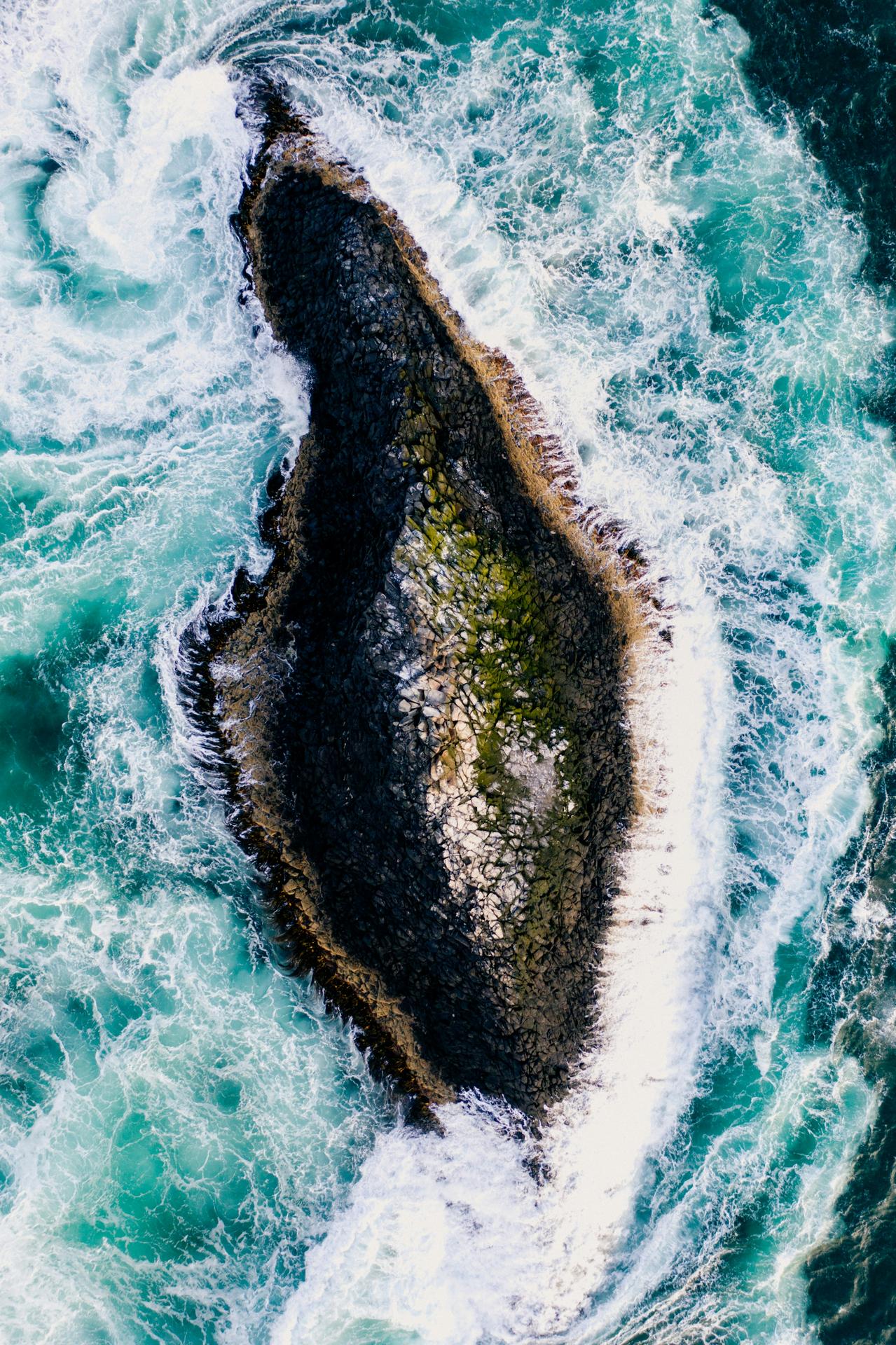 Aerial view of a rocky island surrounded by turquoise ocean waves crashing around it.