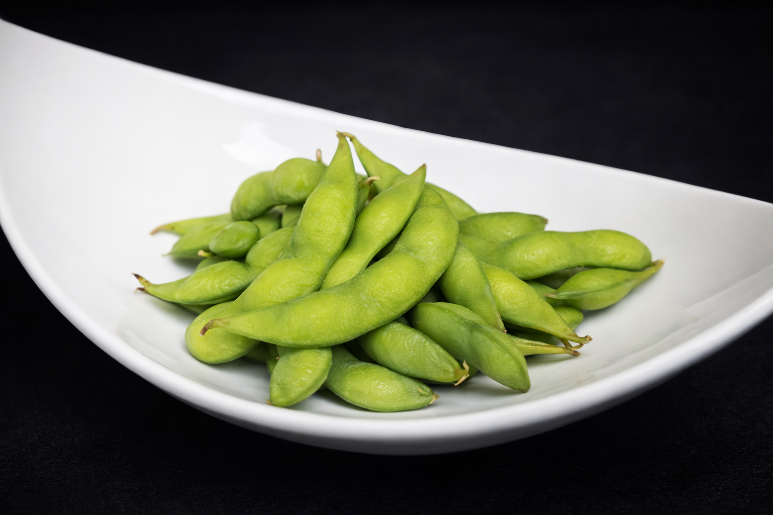 Pile of green edamame beans in pods on a white plate with black background.