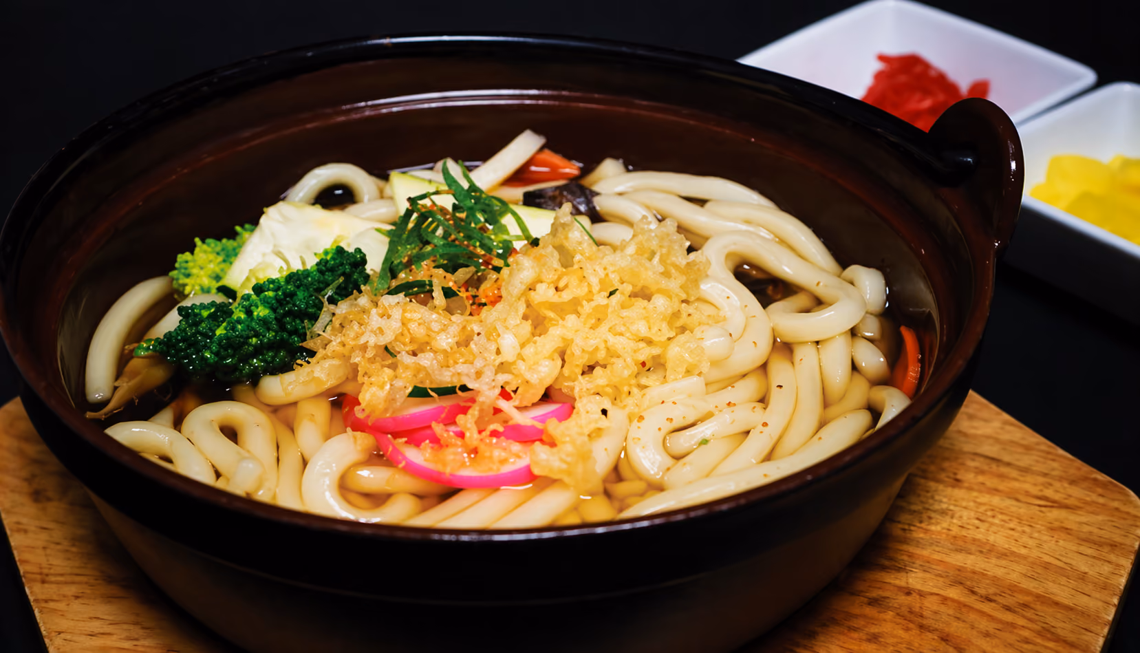 Bowl of udon noodle soup with tempura bits, broccoli, and pink fish cake slices on a wooden board.