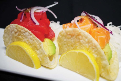 Two sushi tacos with raw fish, avocado, onion slices, and lemon wedges on a white plate.