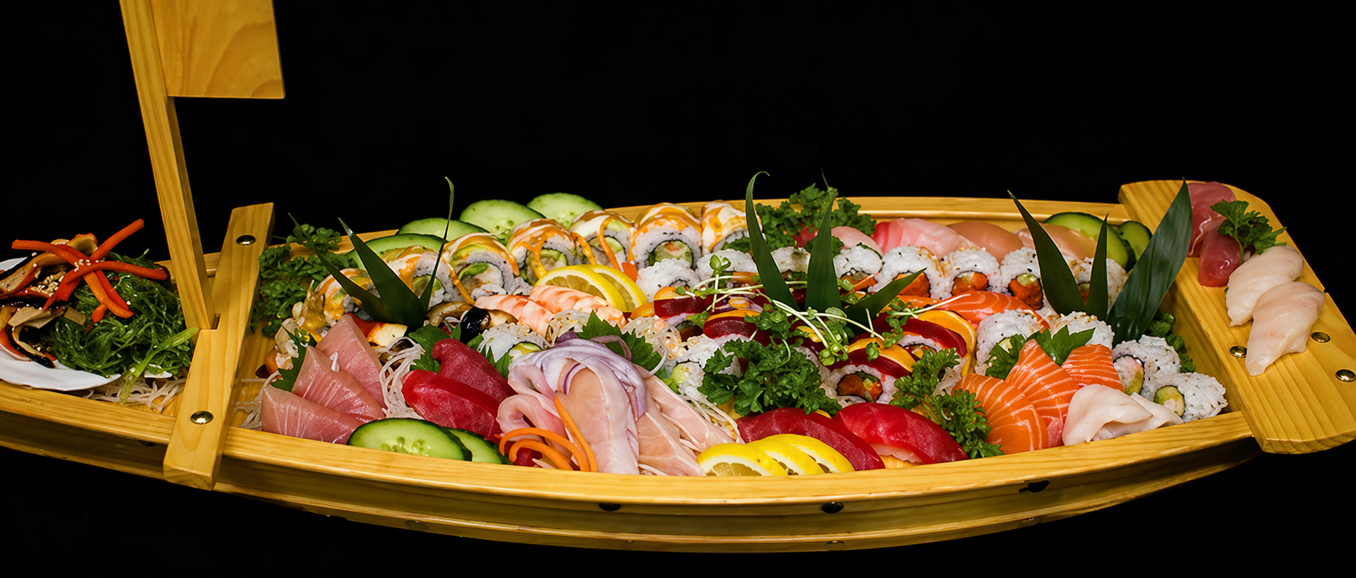 Wooden boat platter with assorted sushi, sashimi, rolls, garnished with cucumber, lemon, and greens.