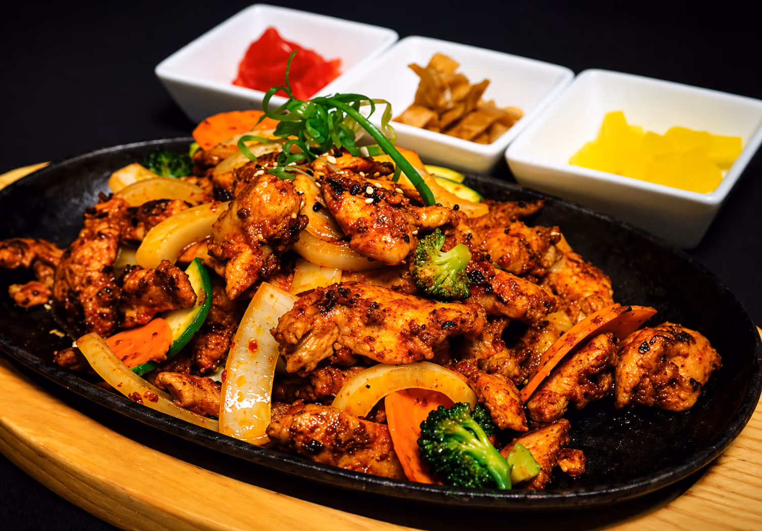Spicy grilled chicken with onions, carrots, broccoli on a hot black plate with three side dishes.