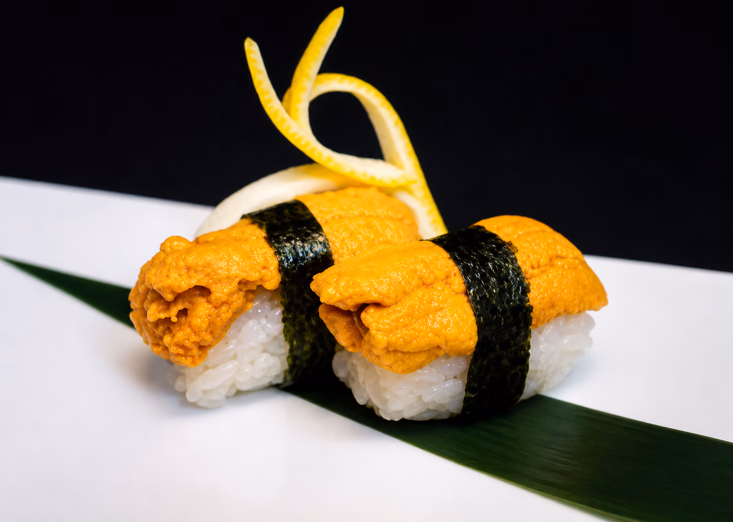 Two pieces of uni (sea urchin) sushi wrapped with nori on rice, garnished with lemon peel on a green leaf.