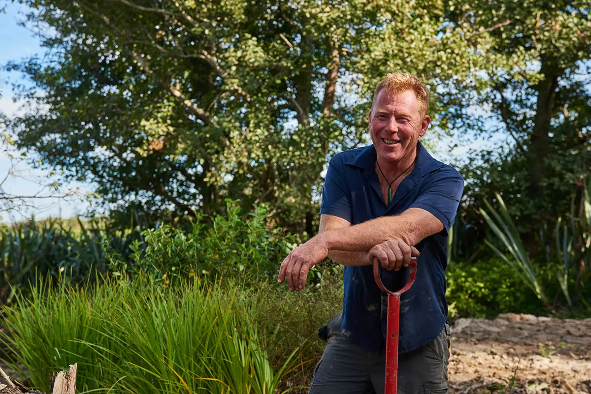 Smiling man leaning on a red garden shovel in a lush outdoor garden setting with green plants and trees.