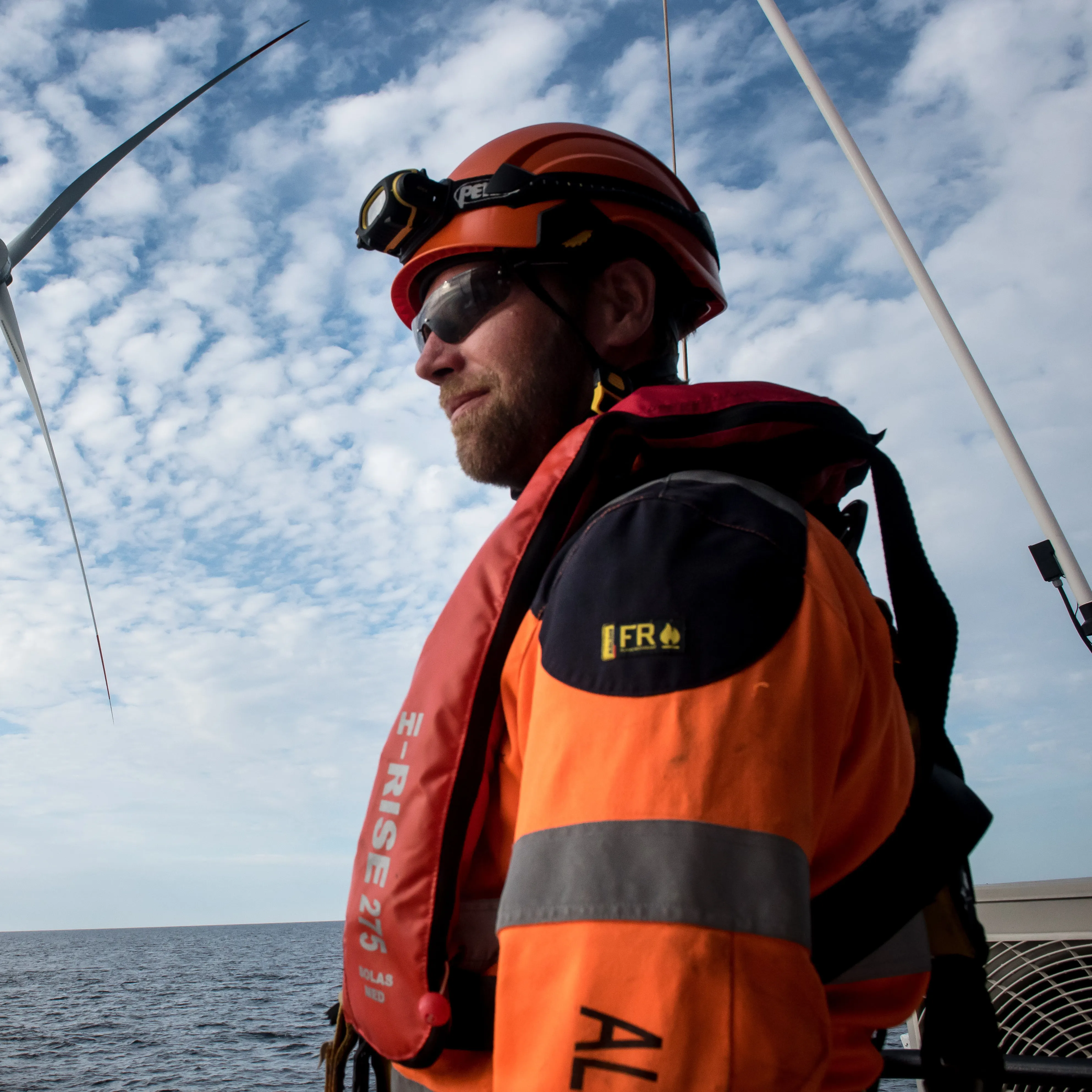 Worker in orange safety gear and helmet with headlamp, standing near offshore wind turbines over the sea.